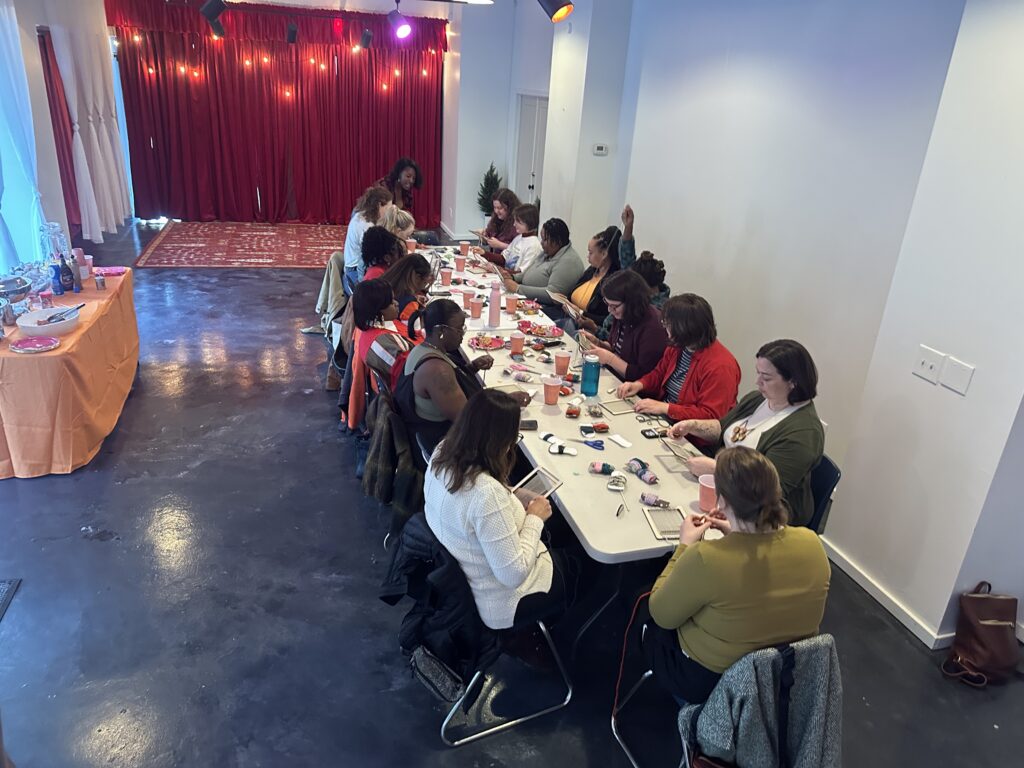 Women sit along long plastic table weaving in a large room with red curtains and a red carpet in the background.