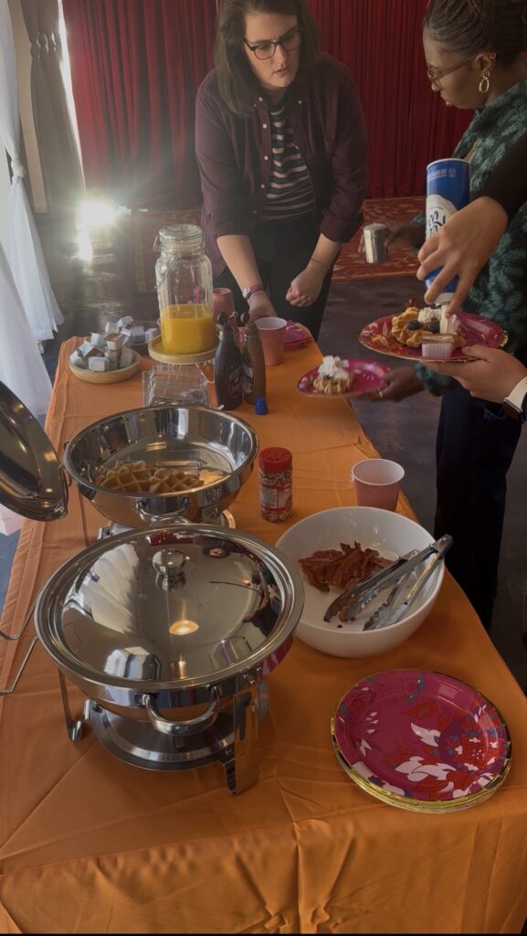 Two women lean over a waffle buffet table while another woman puts whipped cream on a waffle