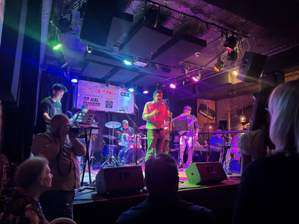 Man talking into mic on night club stage with band setting up on stage behind them.