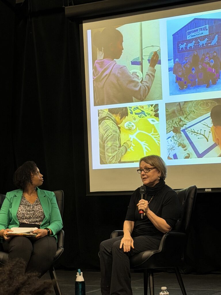 Two women sitting and talking with one of them looking towards a presentation screen.