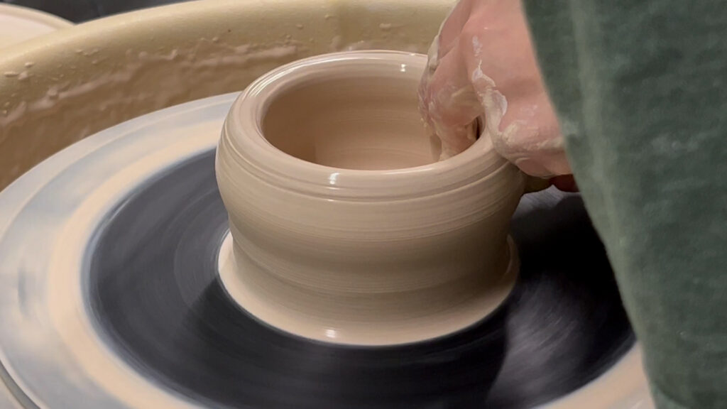 A pot of clay being shaped by hands on a pottery wheel.