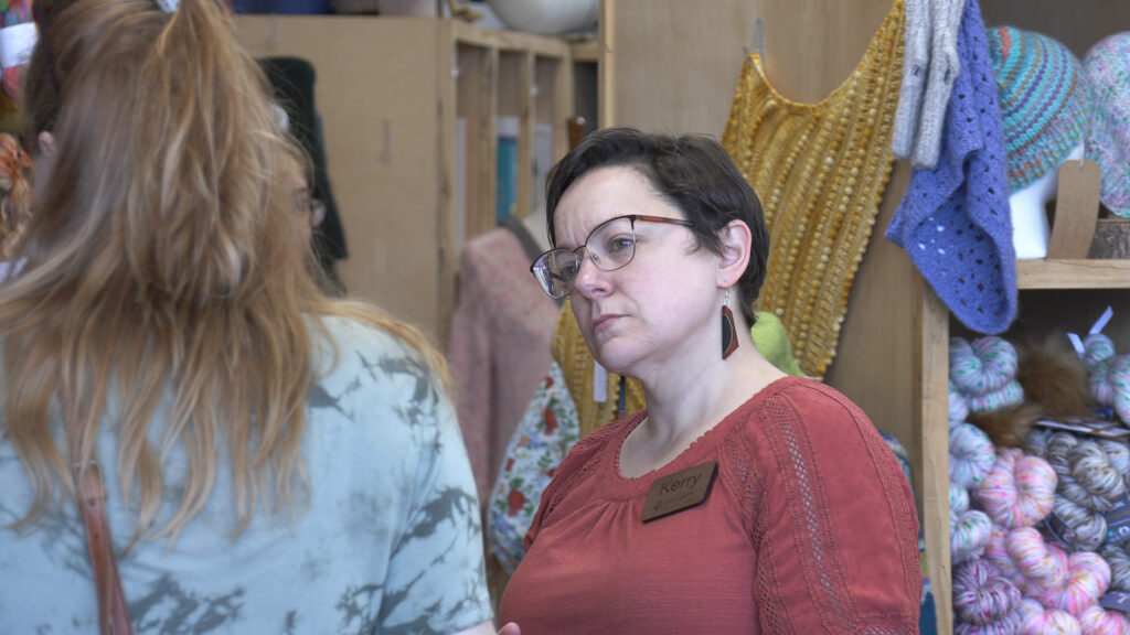 A woman with short dark hair and a name tag reading Kerry speaks with a customer inside a yarn shop, with colorful knitted garments hanging in the background.