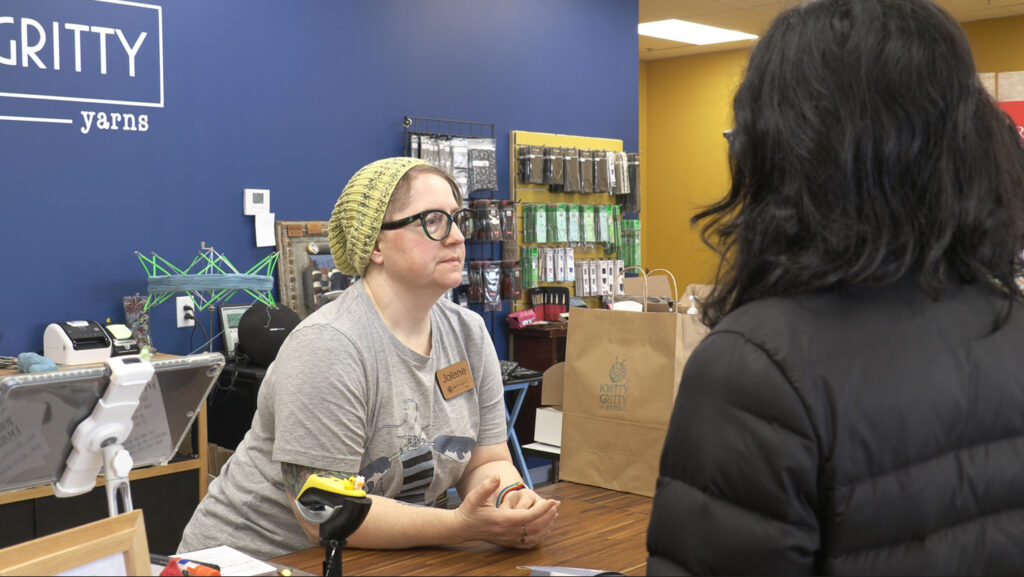 A woman wearing glasses and a knitted hat sits behind a store counter speaking with a customer, with the Knitty Gritty Yarns sign visible in the background.