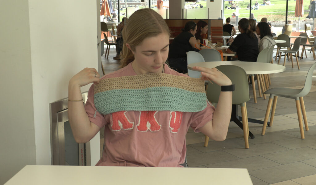 A young woman with blonde hair holds up a partially completed tan and teal crochet panel in a busy campus dining hall.