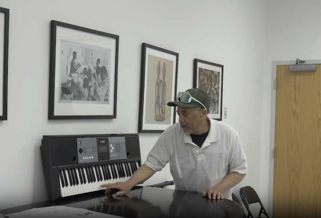 Piano instructor Bernard Wiliford demonstrates notes on a keyboard at the Community Folk Art Center classroom, standing beside a table with sheet music while framed artwork hangs on the wall behind him.