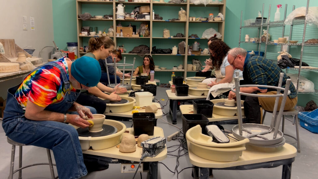 An art studio full of six students work on shaping clay on pottery wheels.