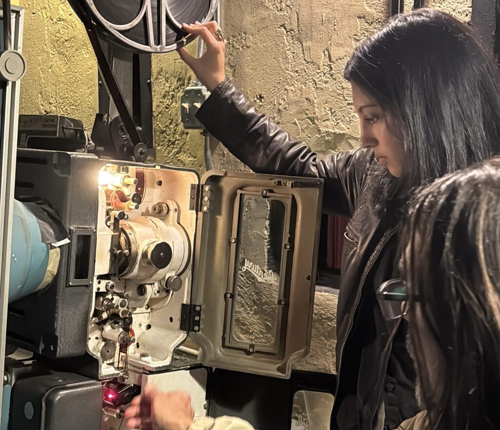 Woman operates a vintage 35mm film projector while another person assists, opening the machine and adjusting the film inside at a theater.