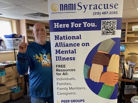 Man standing next to a pull-up banner promoting resource for people wo struggle with mental illness including peer groups, supports groups, education and much more.