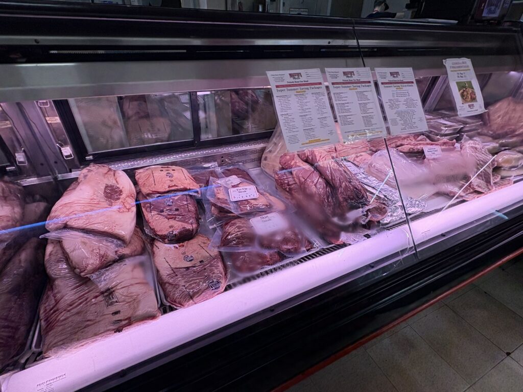 A refrigerated display case containing vacuum‑sealed briskets and other large beef cuts, with printed menus placed above the case.