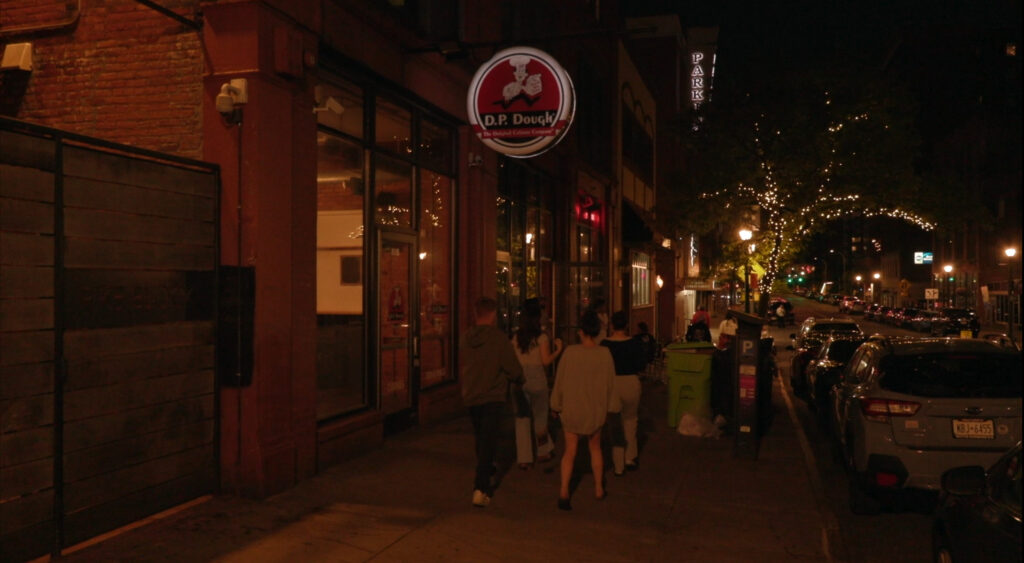 A nighttime street scene in downtown Syracuse showing pedestrians walking past D.P. Dough restaurant, with string lights in trees and storefronts lit along the block.