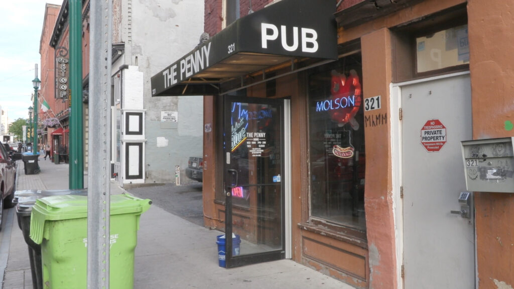 Exterior of The Penny Pub bar in downtown Syracuse, featuring a black awning with white lettering, a neon open sign in the window, and a Molson beer sign visible behind the glass door.