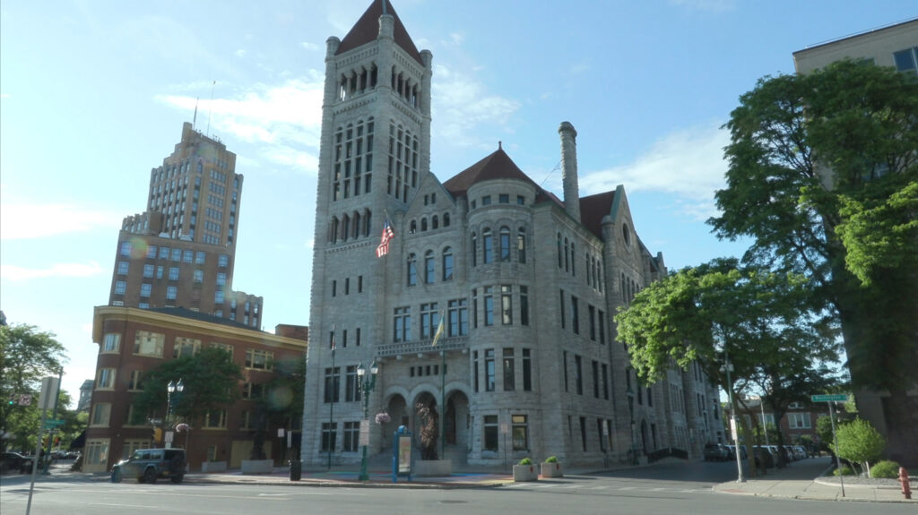 Exterior of Syracuse City Hall, a large gray stone building with a tall clock tower, American flag displayed at the entrance, photographed on a clear sunny day.