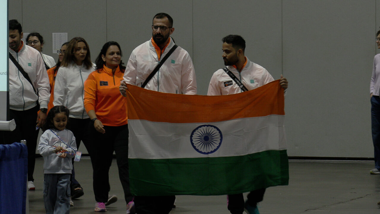 Two men wearing white jackets hold the Indian flag, green stripe on the bottom, white stripe in the middle, orange stripe on top with a blue wheel in the middle of the flag. Behind them is a long line of people including four women, another man, and a child.