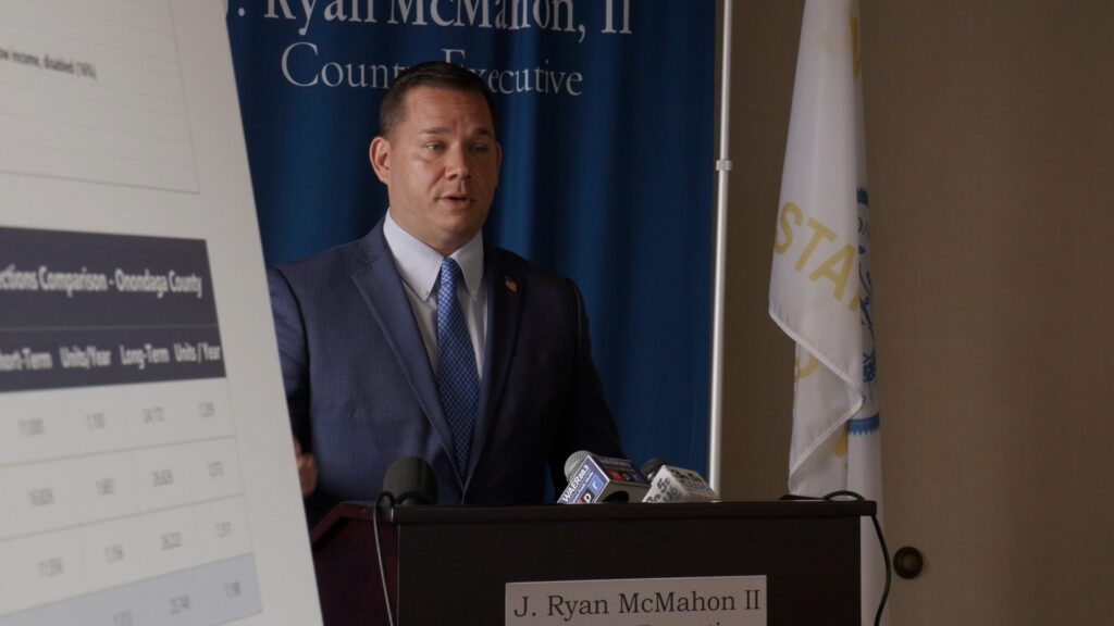 Onondaga County Executive Ryan McMahon stands at a podium with his name on it, mouth open, wearing a navy suit.