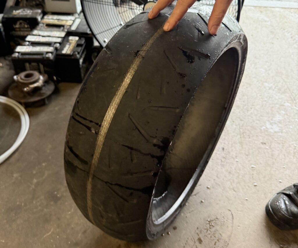 A close-up of a heavily worn motorcycle tire with exposed cords being pointed to inside a repair shop.