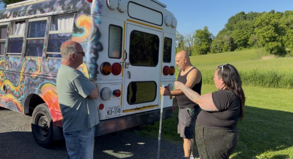 Three people stand behind a brightly painted mini bus covered in colorful artwork while discussing the vehicle in a grassy outdoor area.