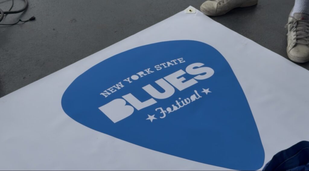 A blue guitar-pick-shaped New York State Blues Festival logo displayed backstage at the festival.