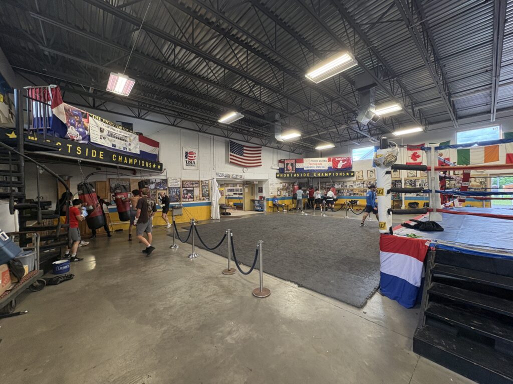 A large, warehouse-style building with a boxing ring on the right hand side. Boxers fill the room training on punching bags.