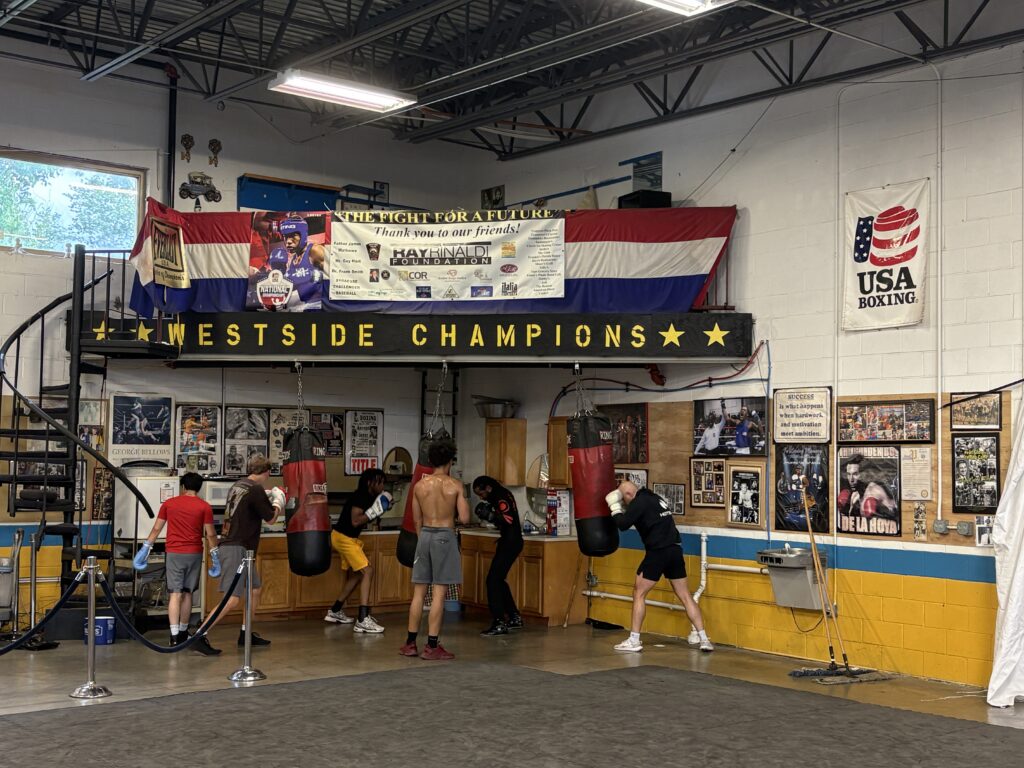 Inside a gym, several boxers train using red punching bags that hang beneath a black balcony that reads "Westside Champions" in yellow text.