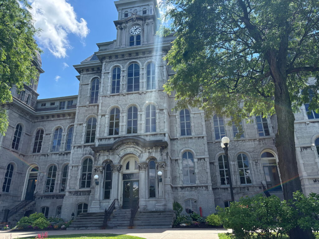 Exterior view of Syracuse University's Hall of Languages, a stone Gothic-style building with a clock tower, framed by green trees against a blue sky.