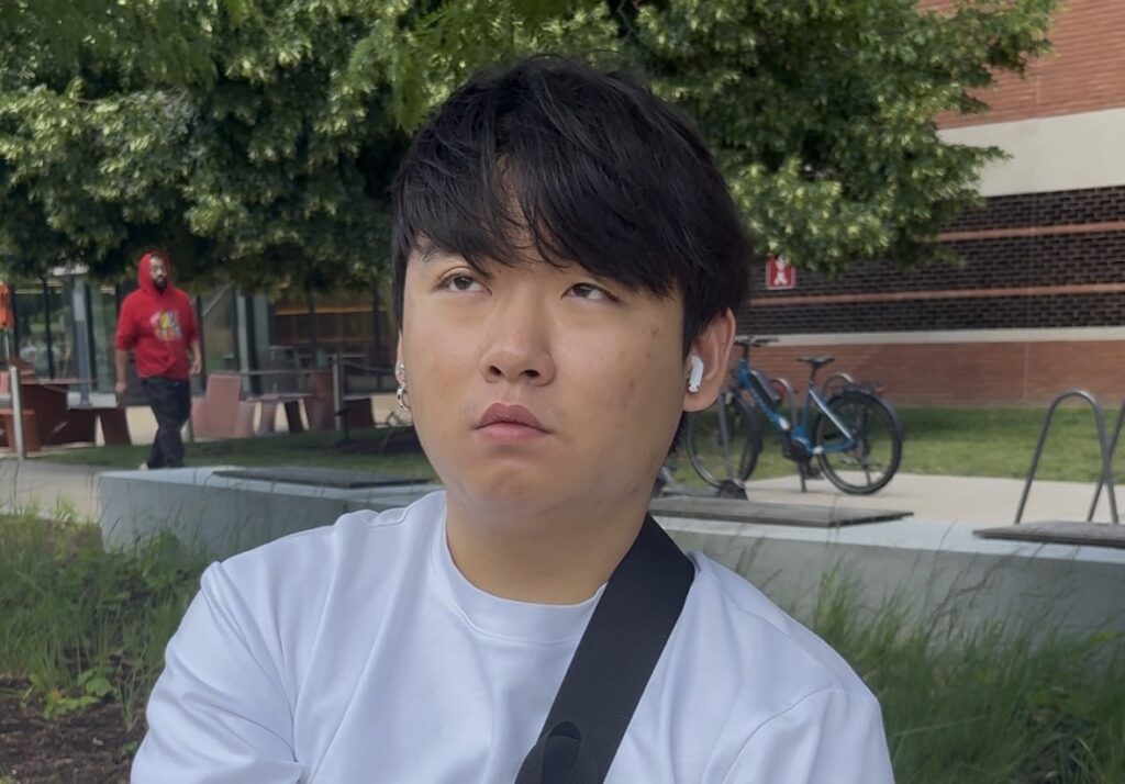 Aidan Jeong sits outside on a campus bench, looking off to the side, with a bicycle rack and brick building in the background.