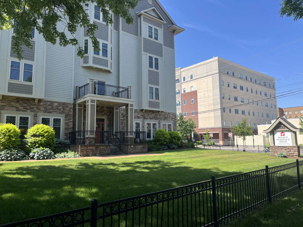 The white and brown Ronald McDonald House sits on a green lawn with a black fence in front.