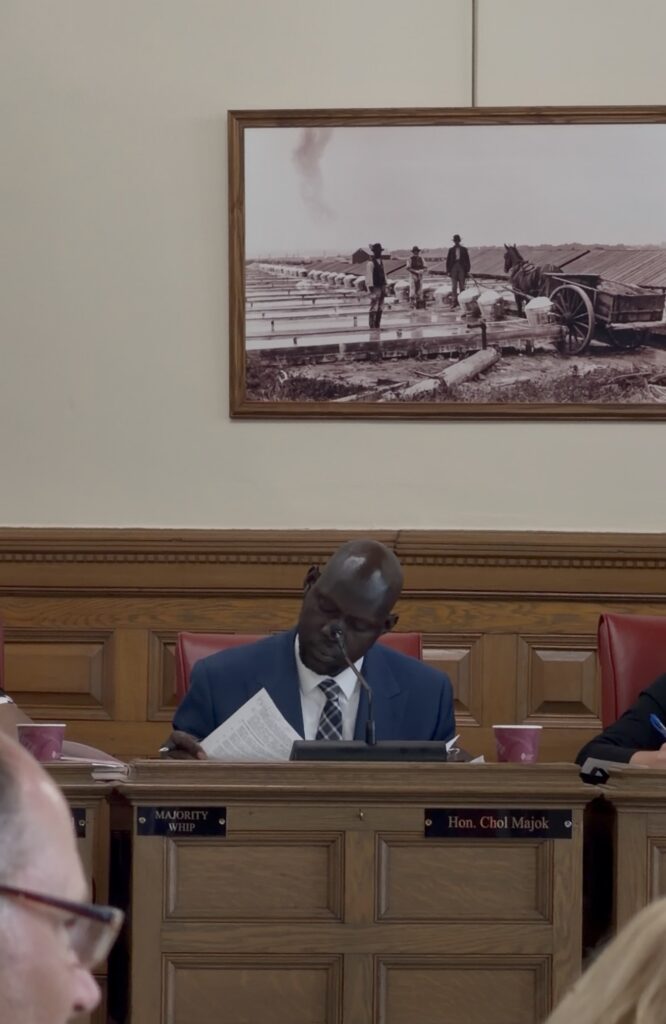 A man sitting at desk with a name plate on the front - Hon. Chol Majok - reading a piece of paper