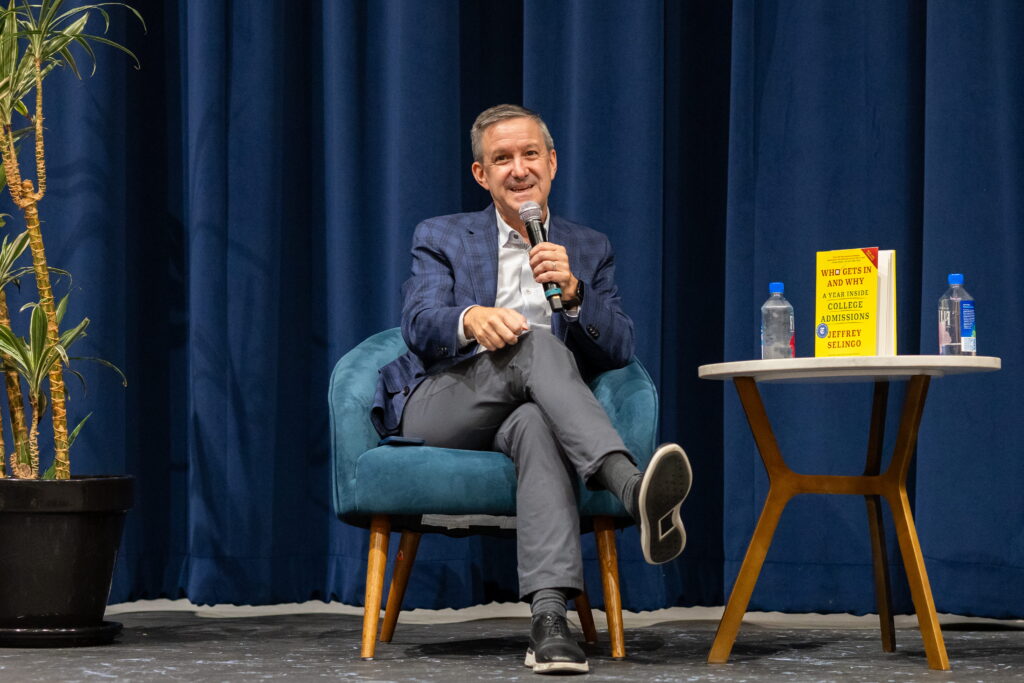 Jeff Selingo sits in a teal armchair on stage holding a microphone during a fireside chat, with his book "Who Gets In and Why" displayed on a table beside him.