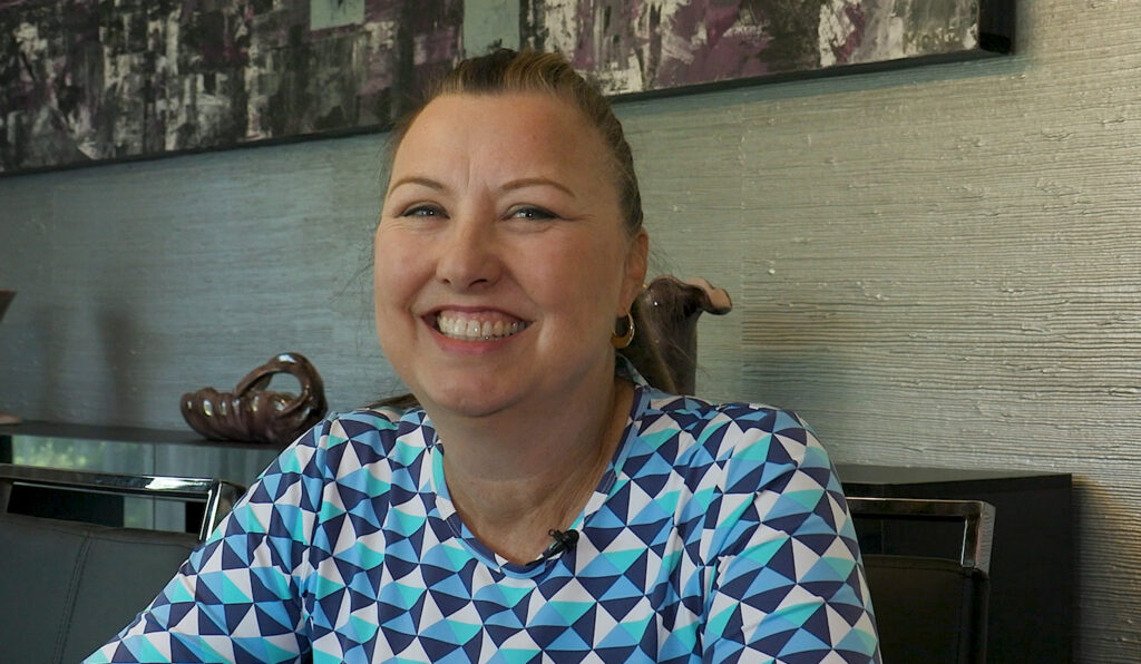 A woman smiling in a dining room wearing a blue patterned shirt.