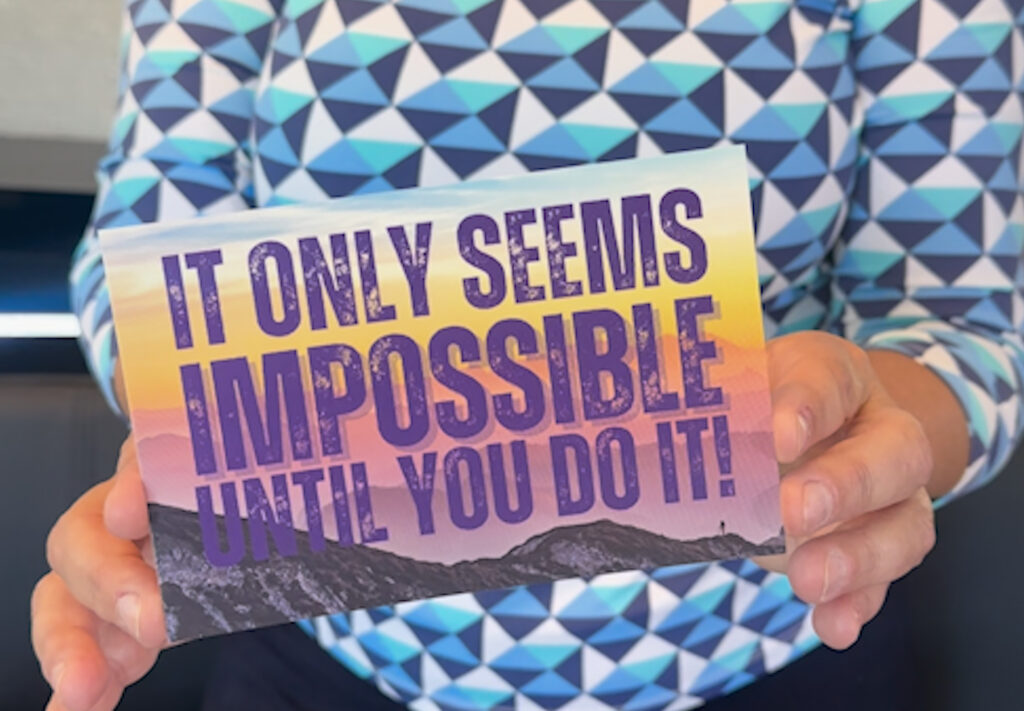 A blue patterned shirt with two hands holding a postcard thats yellow and purple with words saying, "it only seems impossible until you do it."