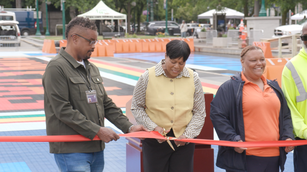 Mayor Sharon Owens cuts a red ribbon with gold scissors as city staff looks on. Behind them, the rink's modular floor runs in stripes of red, orange, yellow, green and blue, lined by orange barriers.