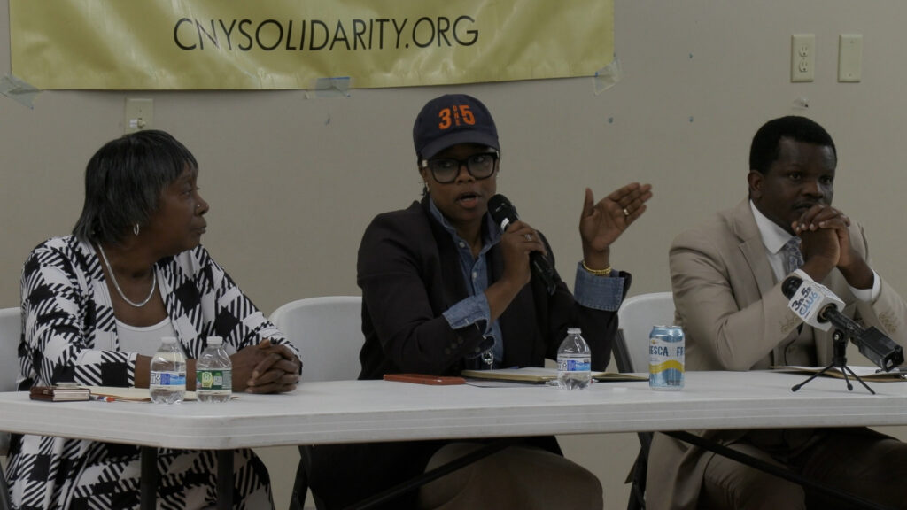 Two women and one man sit next to eachother at a folding table. The woman in the center is speaking and holding a microphone.