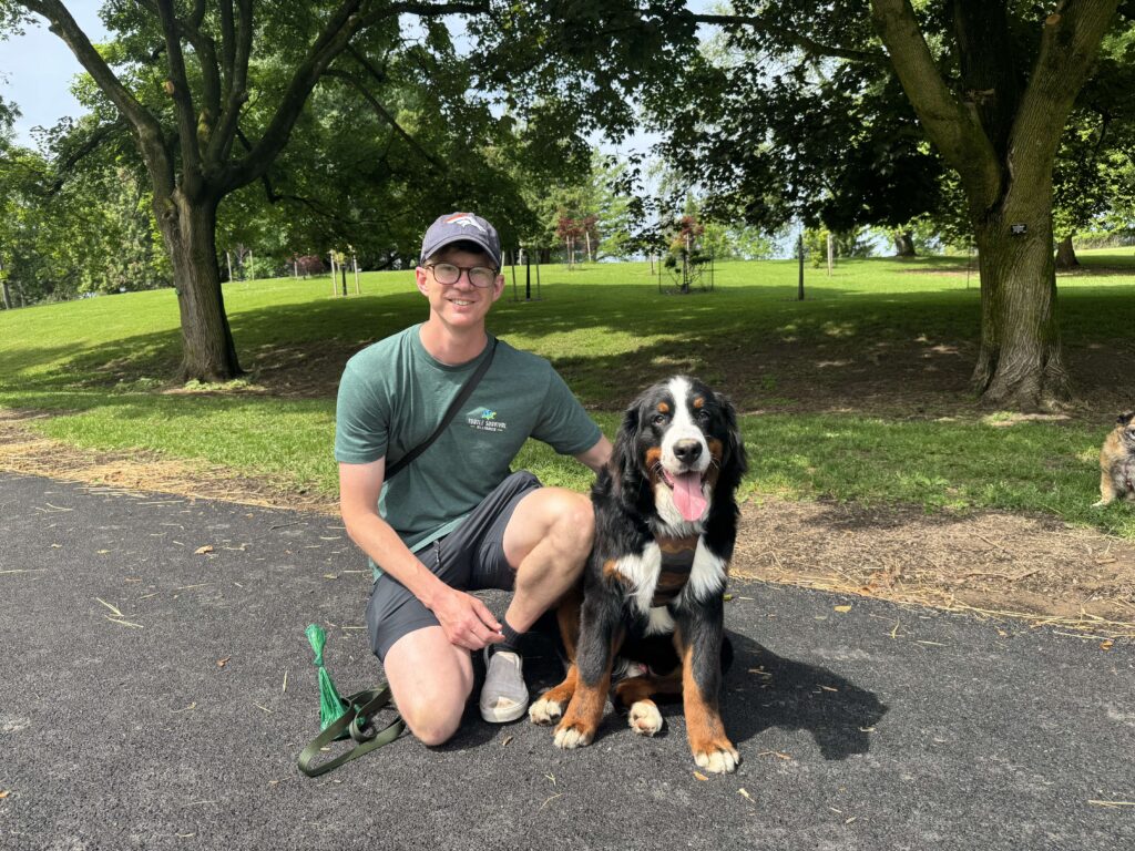 Man kneels with dog on path with trees and grass behind them