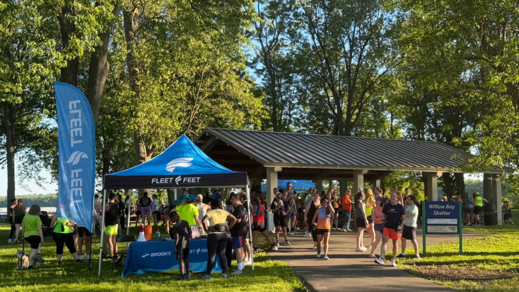 Large group of people meeting outside under a pavilion. A fleet feet tent is off to the left