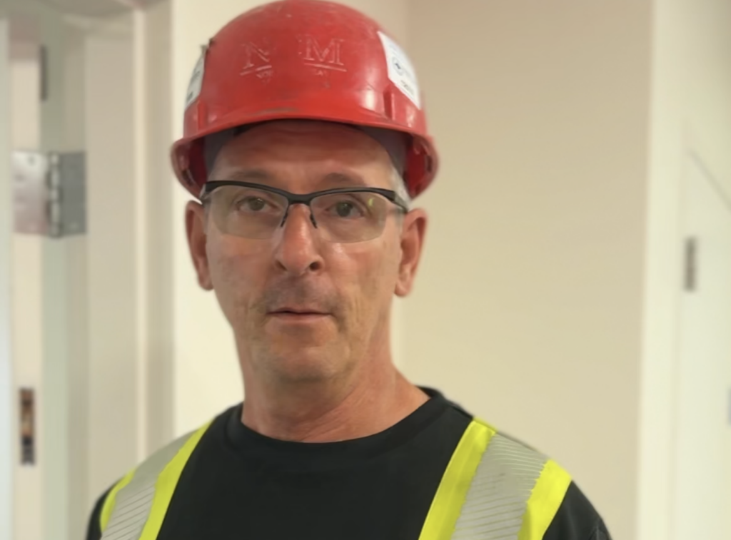 Construction worker in a red hard hat, glasses, black T-shirt and a neon vest.