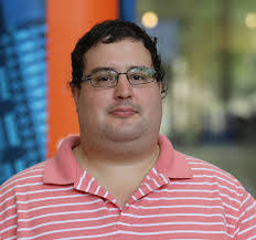 Headshot of Jason Pollack, a man with curly dark hair and glasses, wearing a pink striped polo shirt, against a blurred background.