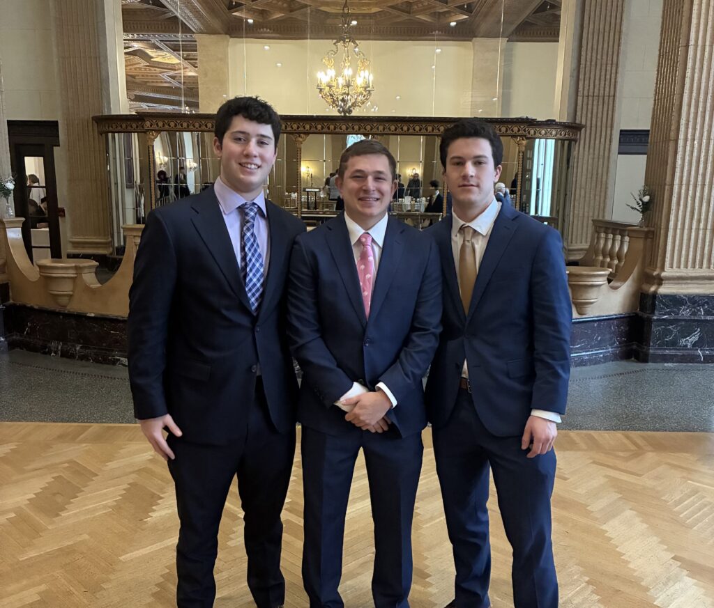 Three young men in navy blue suits standing together for a photo, smiling for the camera inside of a nice ballroom.