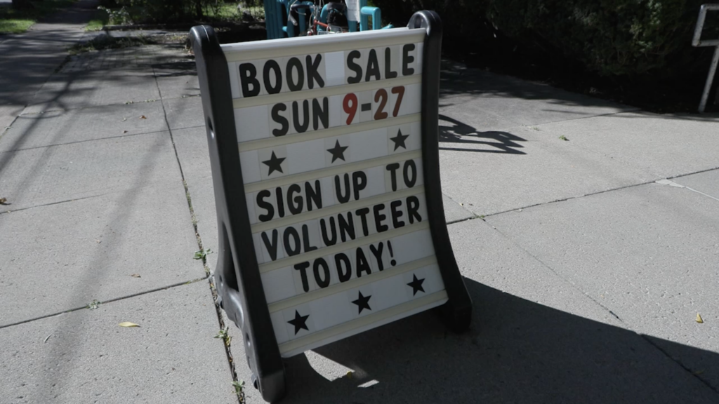 A sign advertising a book sale on Sunday, September 27 and telling volunteers to sign up today sits on a concrete sidewalk.