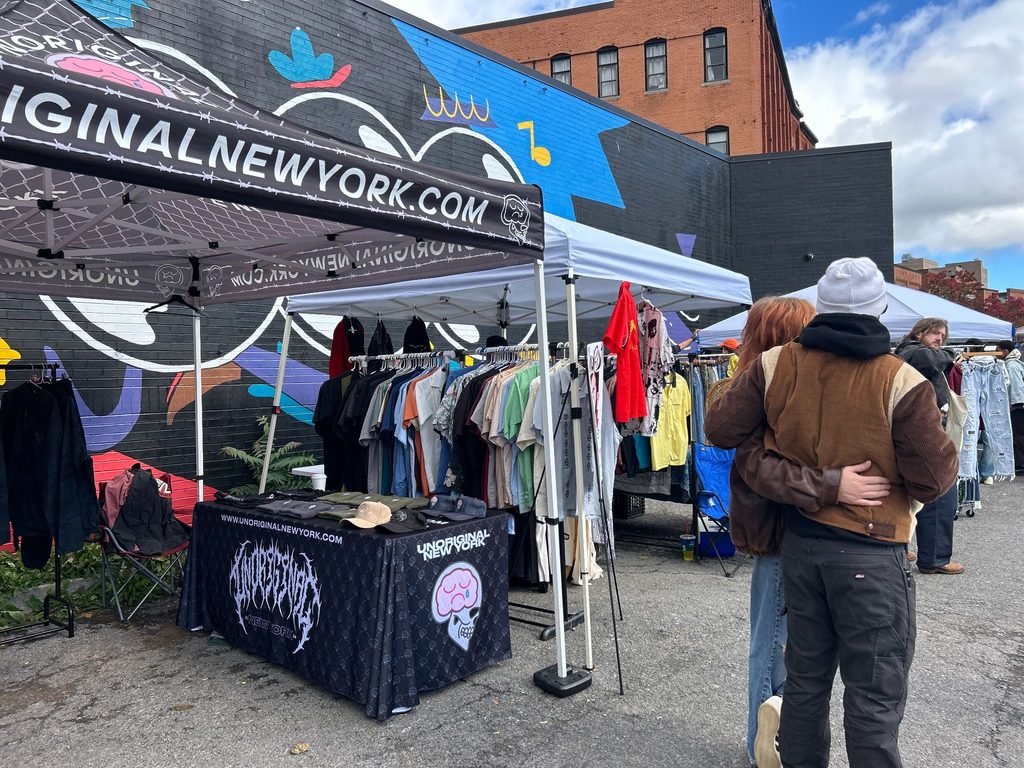Unoriginal New York’s booth, with hats on a table and racks of vintage shirts, sits in front of Sound Garden's black-brick mural as couple, Anna Kilmartin and Vlad, wrap their arms around each other.