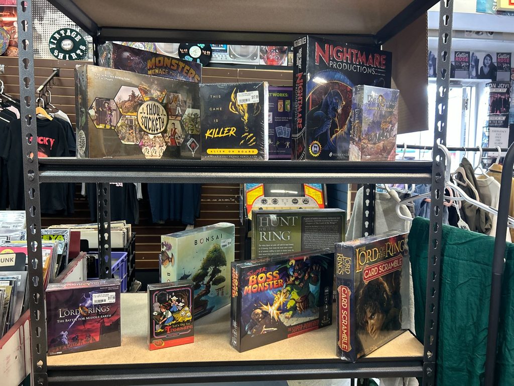 Boxed board games and card games line two shelves in The Sound Garden, with vinyl records and T-shirts behind them.