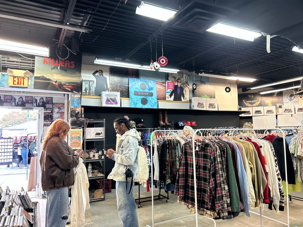 Shoppers stand near Sweet Teeth's racks of vintage flannels and button-up shirts inside The Sound Garden, beneath a wall of album posters and turntables.